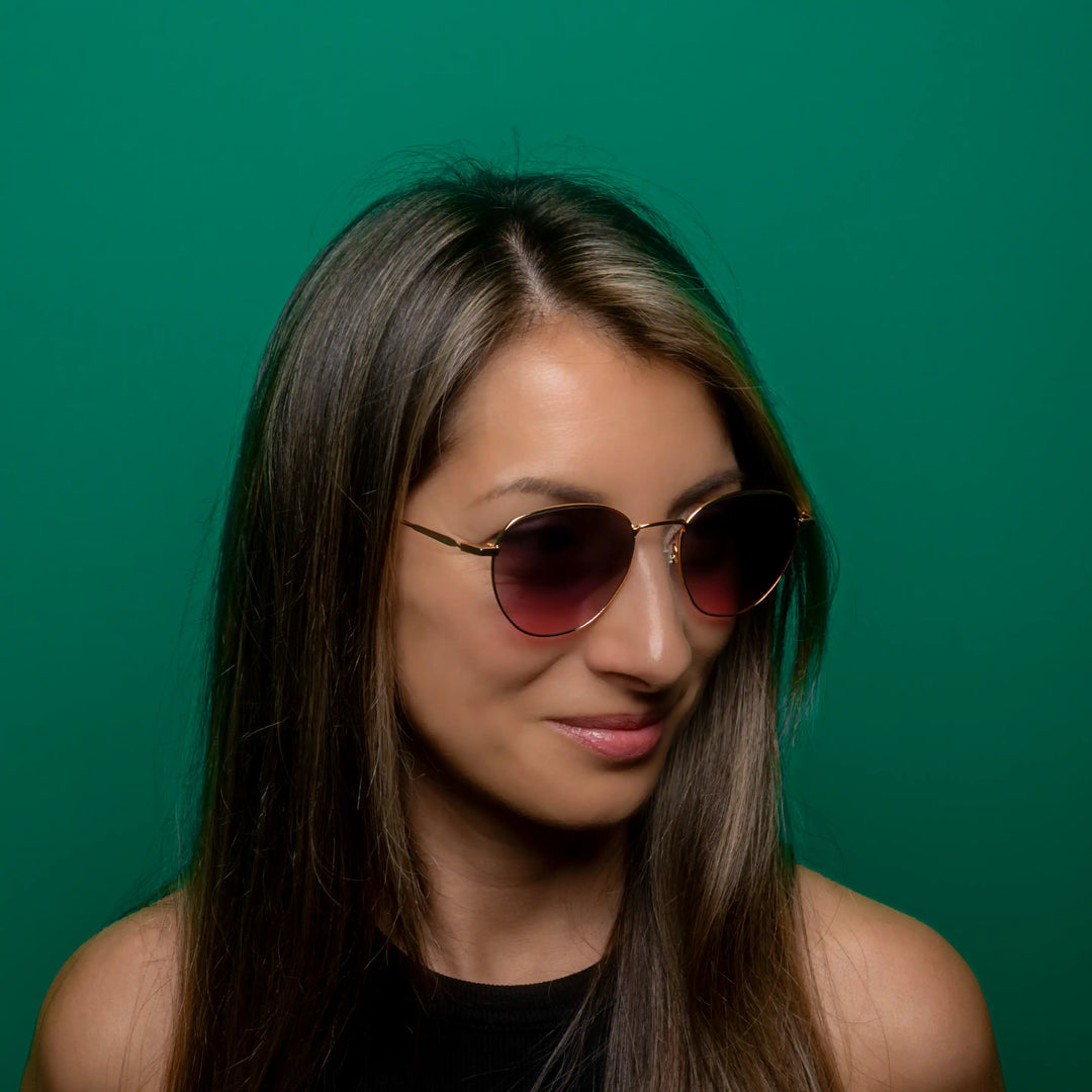 Woman wearing gold colored sunglasses with pink lenses against a green background #desktop-half for half-width