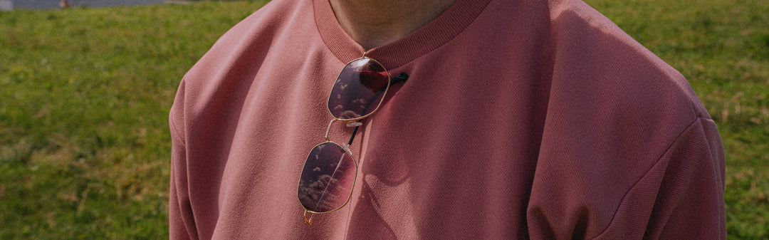 Person wearing a pink sweatshirt with sunglasses hanging from the neckline, standing in a grassy area.